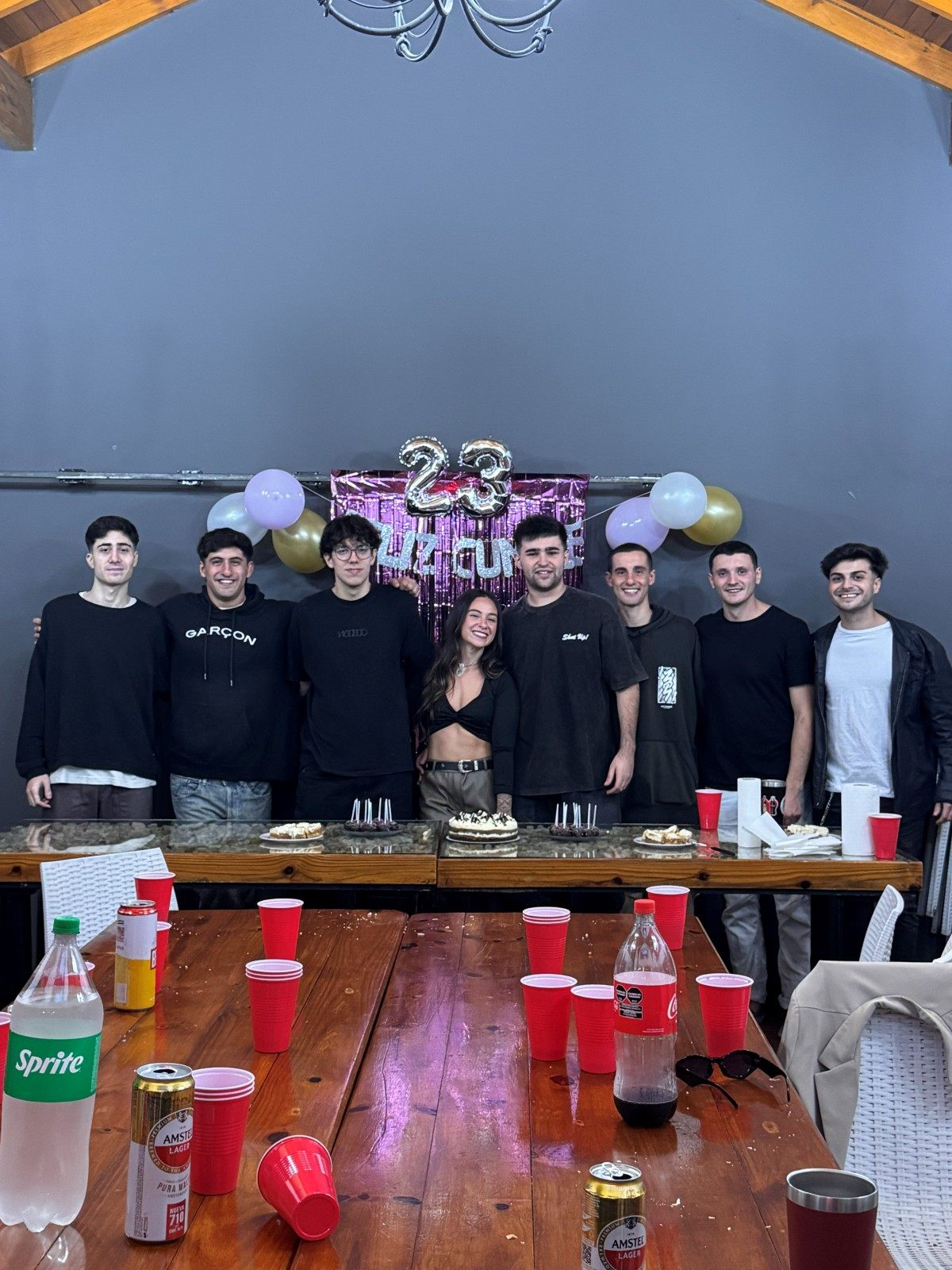 Un grupo de amigos posando detrás de la mesa en un cumpleaños de 23, con globos y cortina metalizada