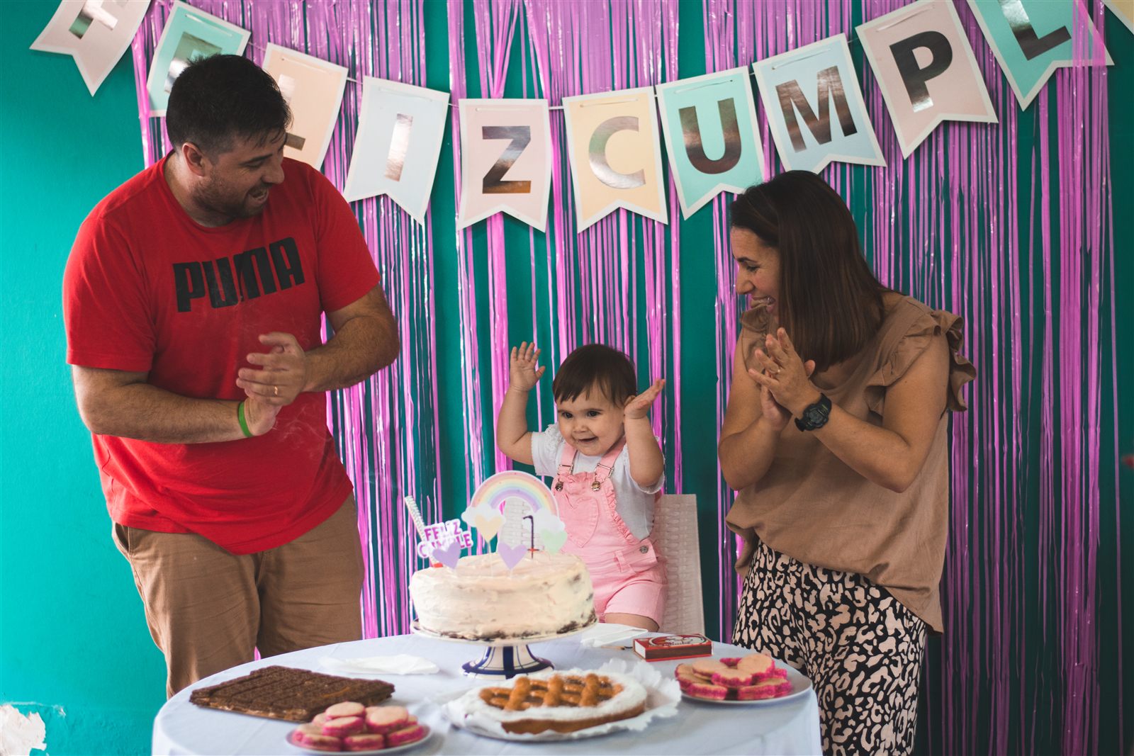 Una beba festejando su primer año frente a la torta, con su mamá y su papá aplaudiendo