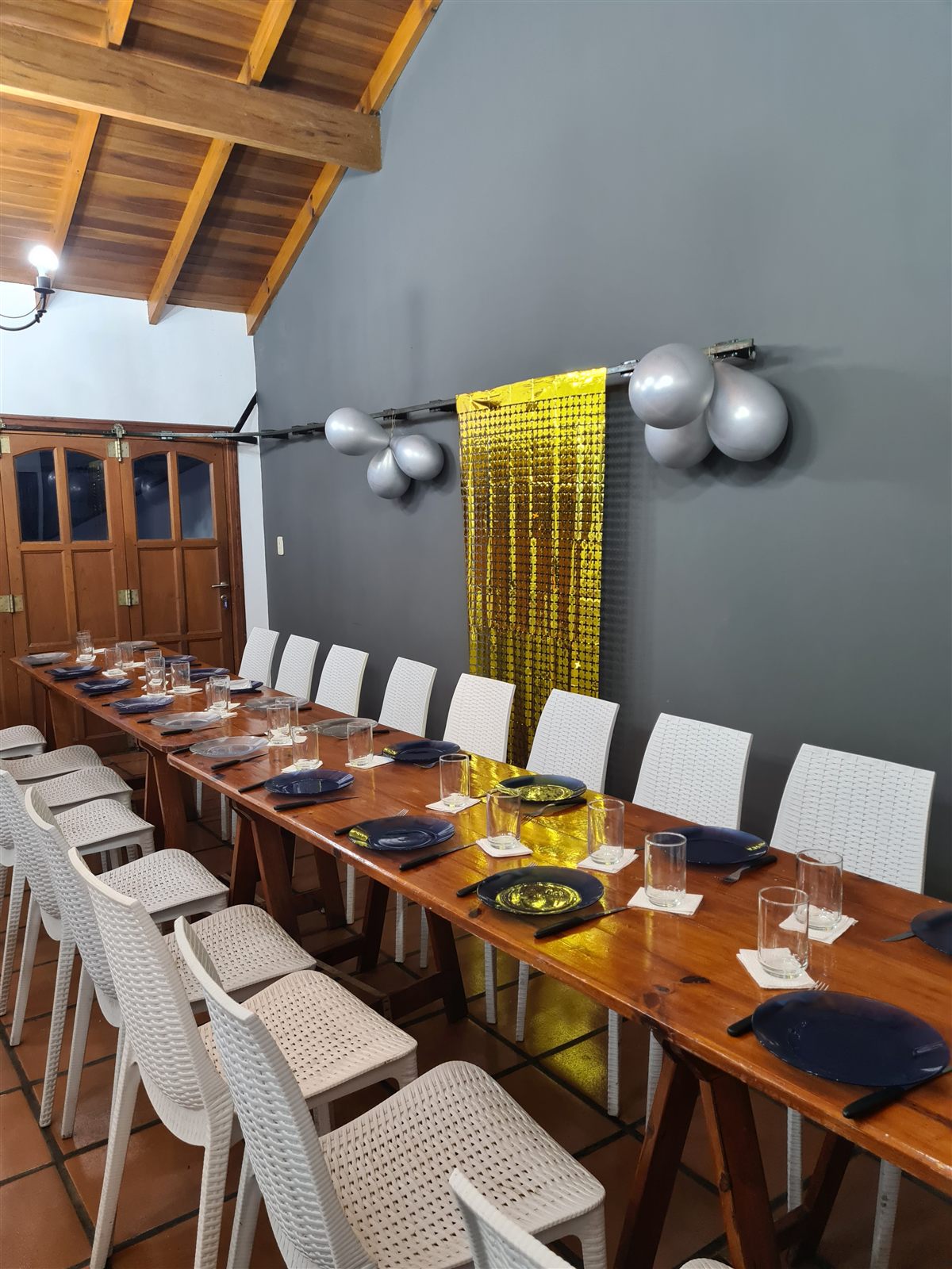 Mesa larga con platos azules, vasos y cubiertos, y una cortina dorada con globos plateados en la pared