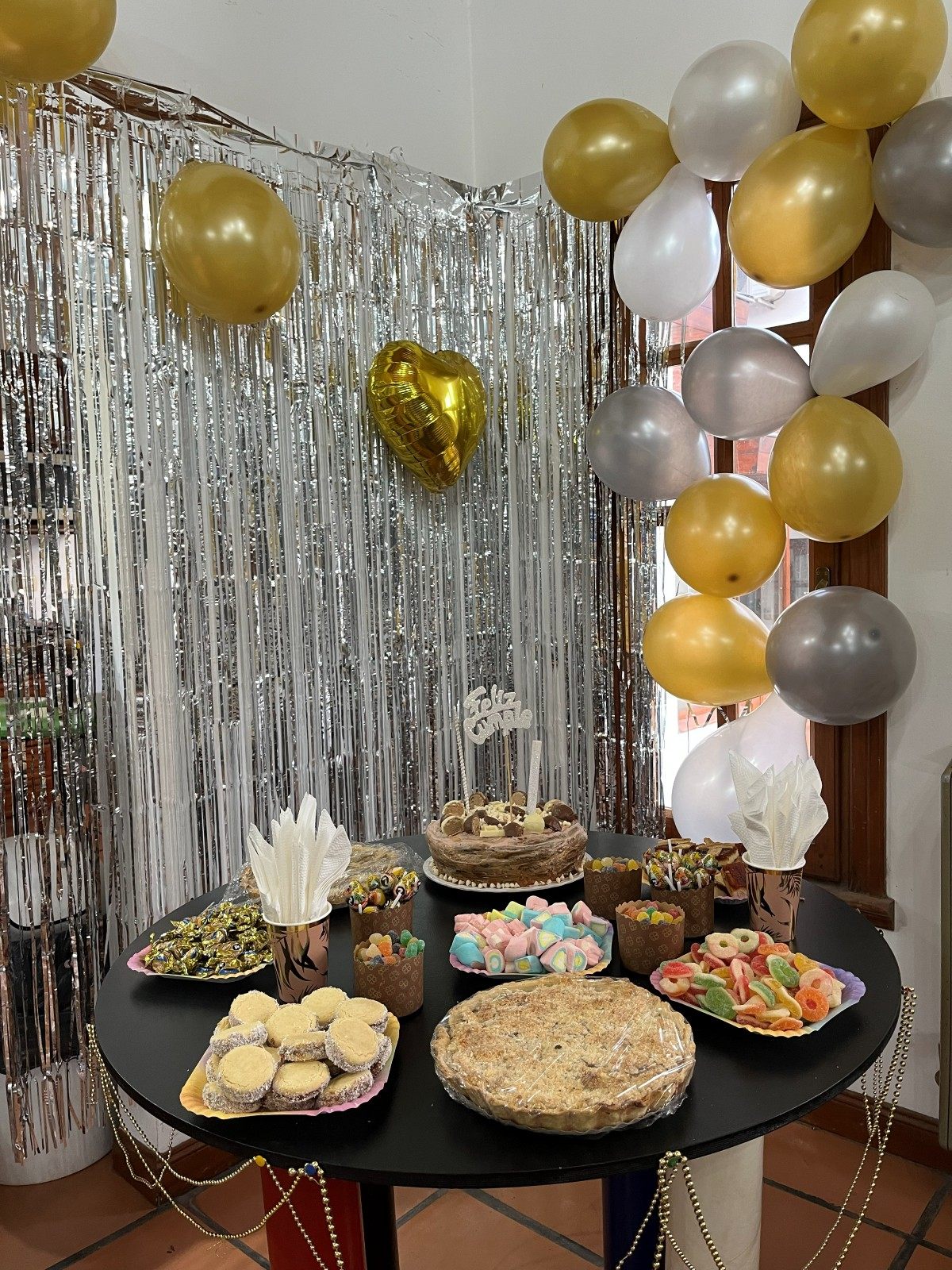 Mesa dulce con torta, alfajores y golosinas, con cortina plateada y globos dorados y blancos