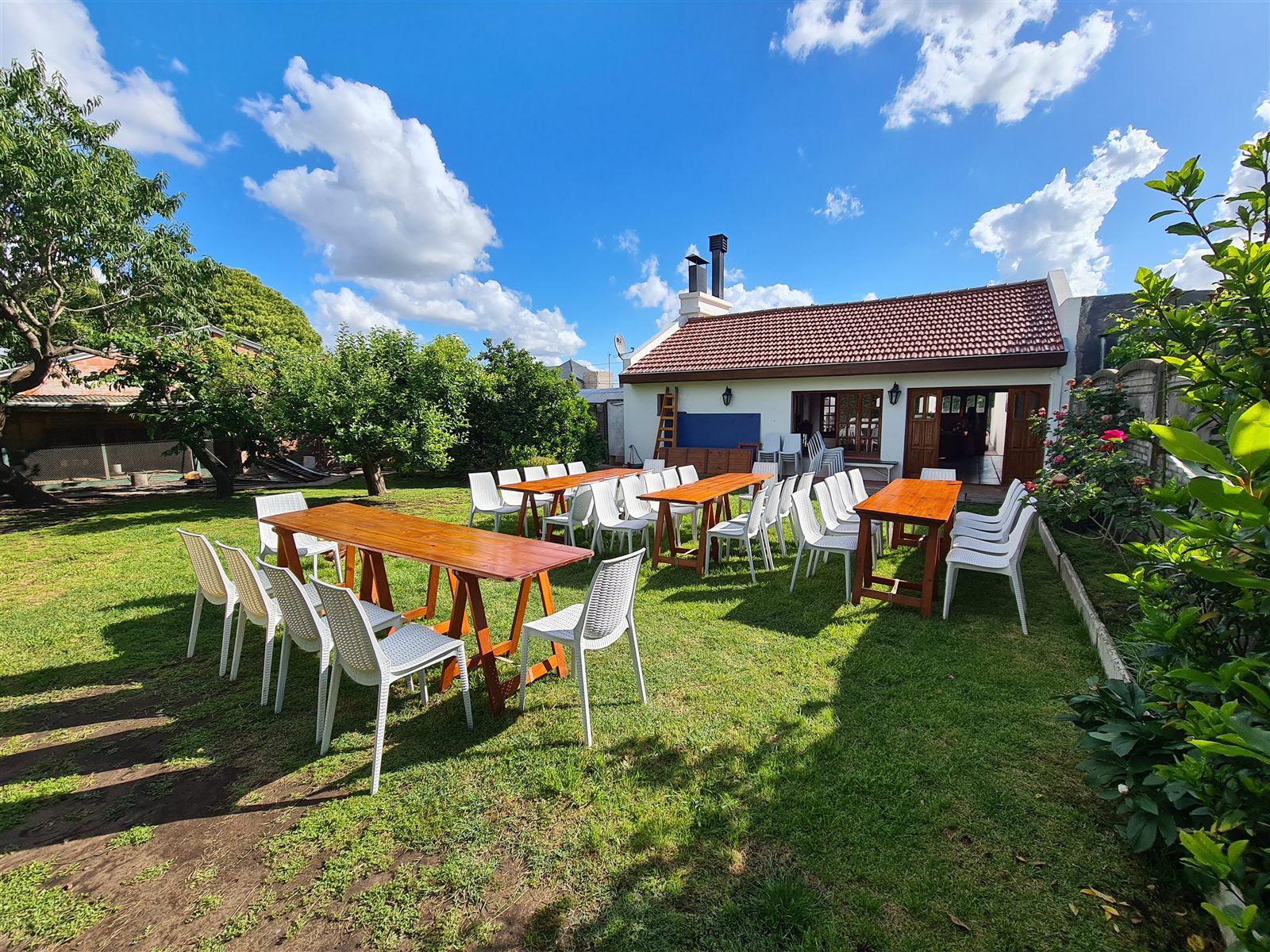 El patio con mesas largas de madera y sillas blancas sobre el pasto, con el quincho de tejas al fondo y cielo despejado