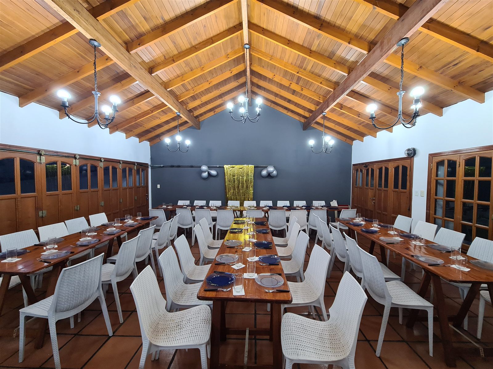 El salón armado para una cena: tres filas de mesas con vajilla, sillas blancas, techo de madera con arañas y pared gris decorada