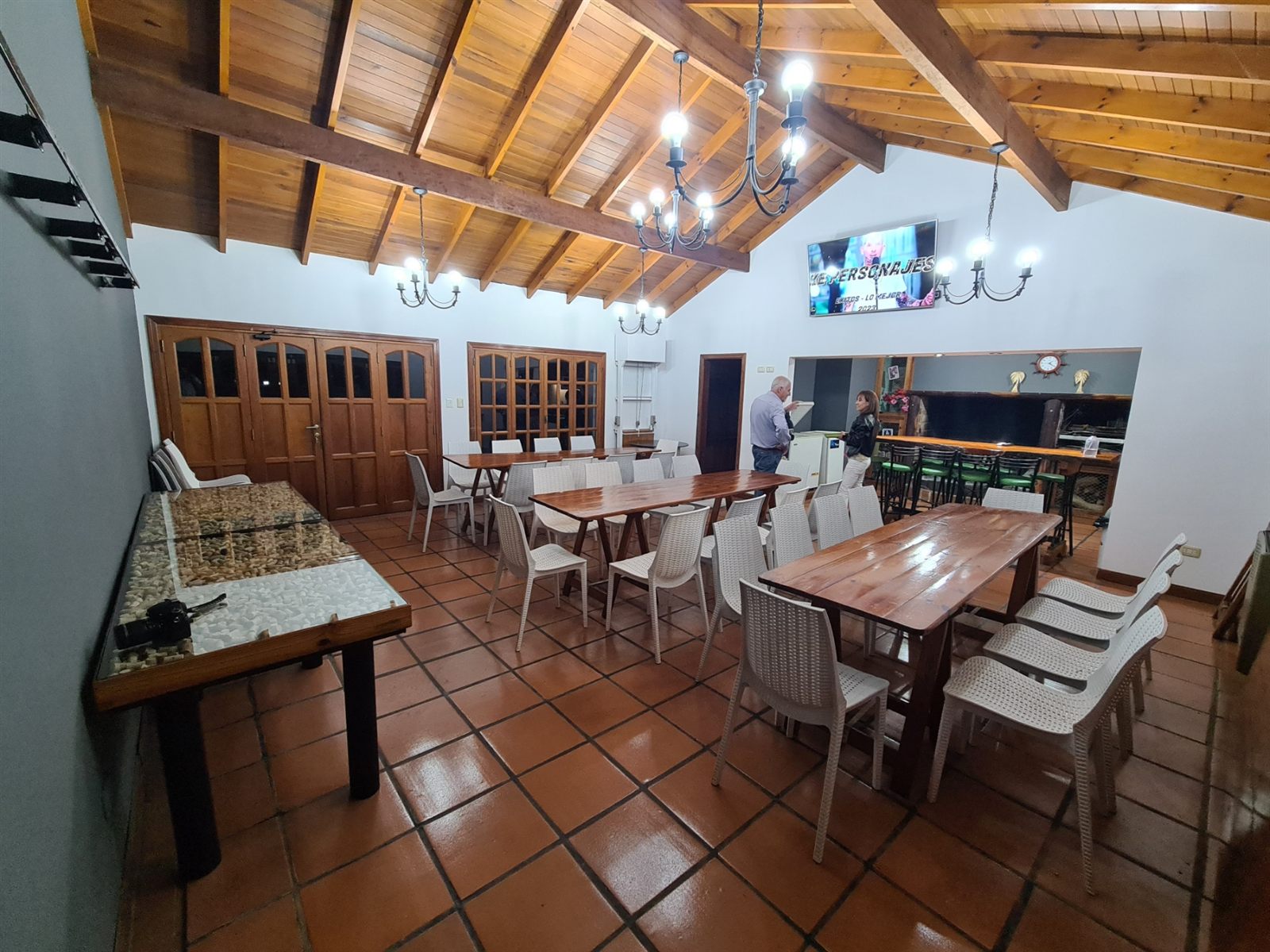 El salón de noche con las mesas, la tele grande, y al fondo la barra de madera con banquetas y la parrilla