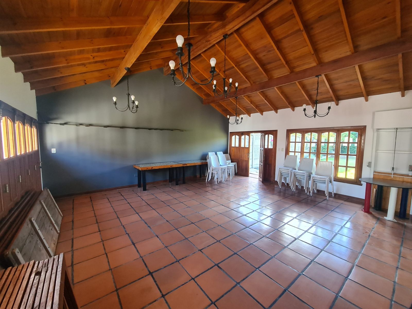 El salón vacío de día: piso de baldosas, techo de madera a dos aguas y ventanales al patio